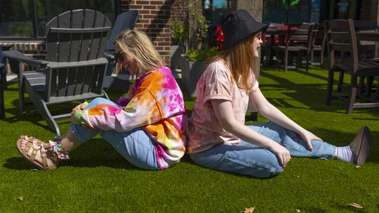 Two young girls wearing tie dye shirts sitting back to back on green grass