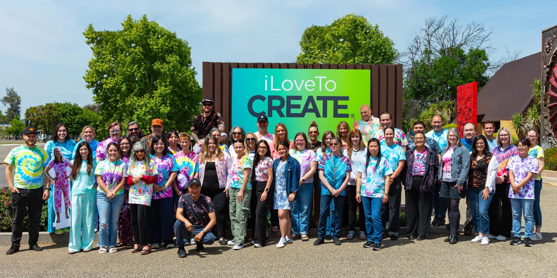 Large group of people posing in front of a 'I Love to Create' sign outdoors.